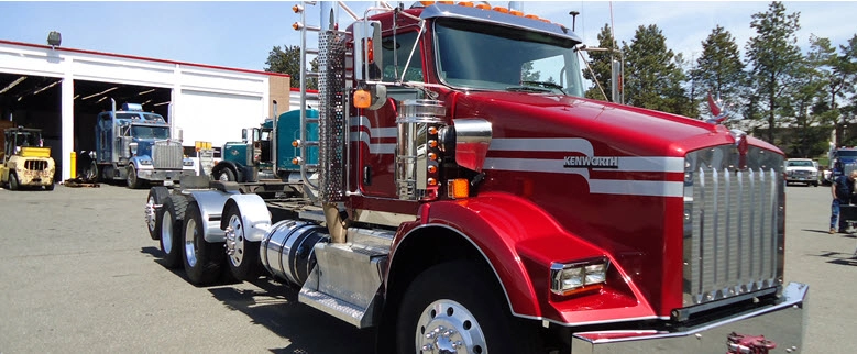 Red Kenworth day cab semi tractor parked in a dealership lot, with other trucks and a service shop visible in the background