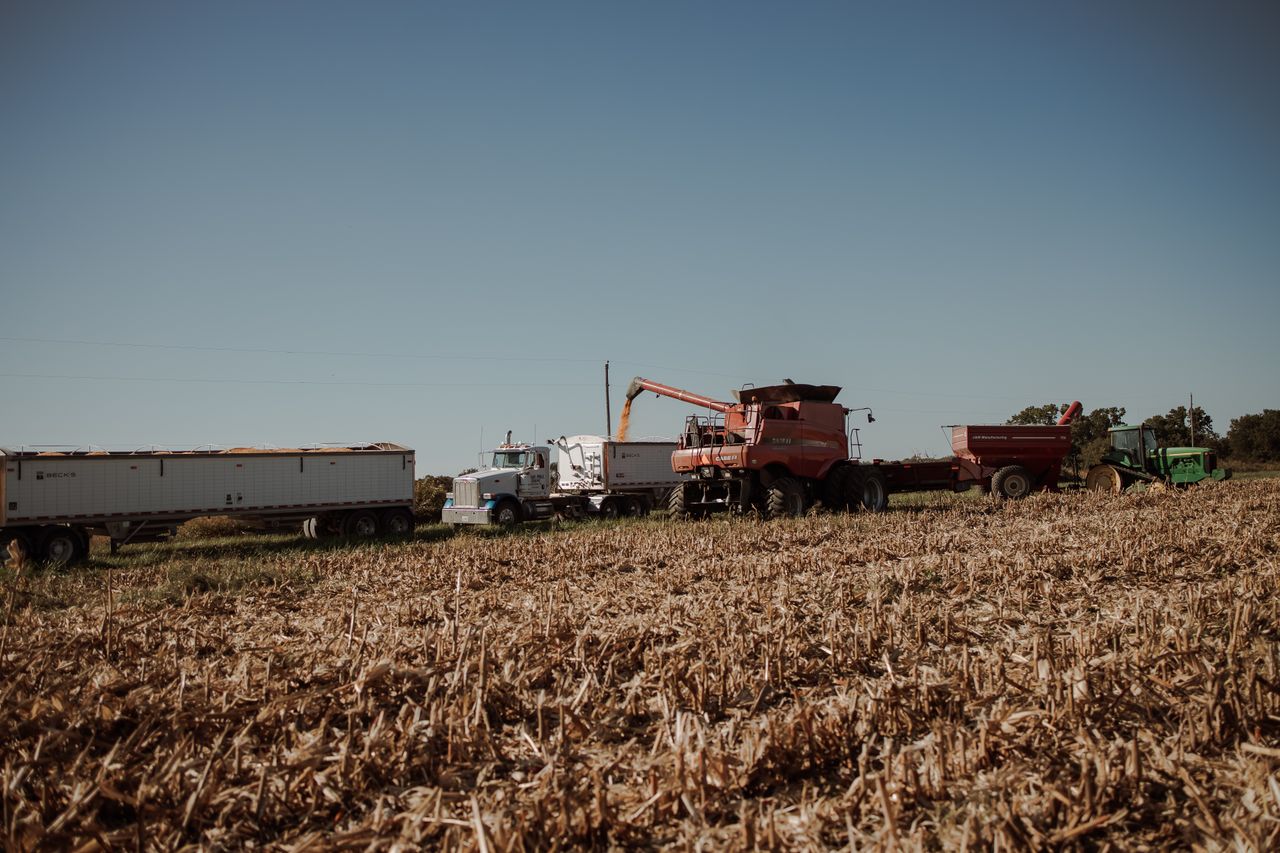 Red combine harvester unloading grain into a grain cart beside a green tractor and a white semi-truck with grain trailers in a harvested field under a clear blue sky