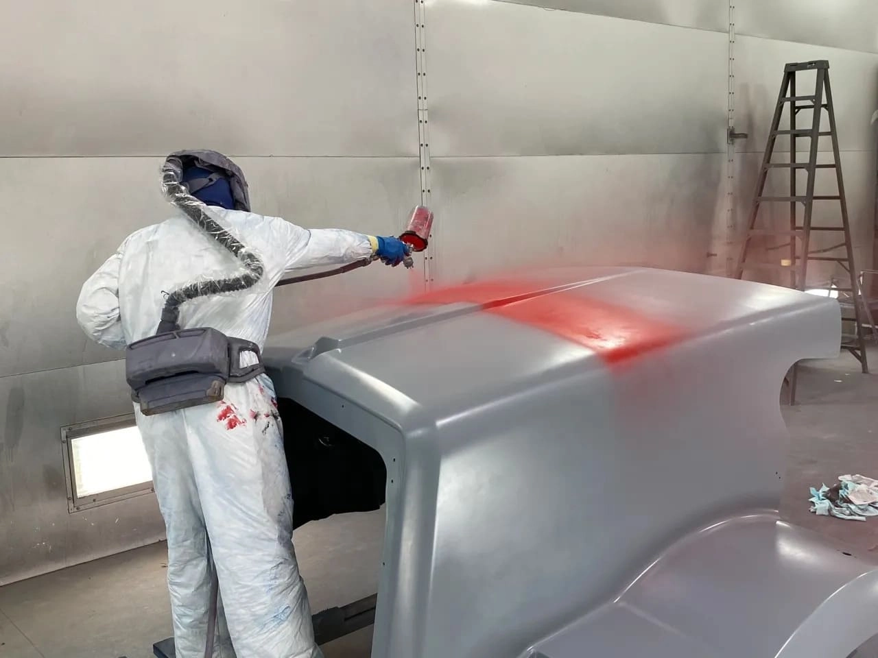 A technician in a white protective coverall and supplied-air respirator hood uses a spray gun to apply red paint over a primed gray truck hood/cab shell inside a metal-walled downdraft paint booth, with a ladder visible in the background.