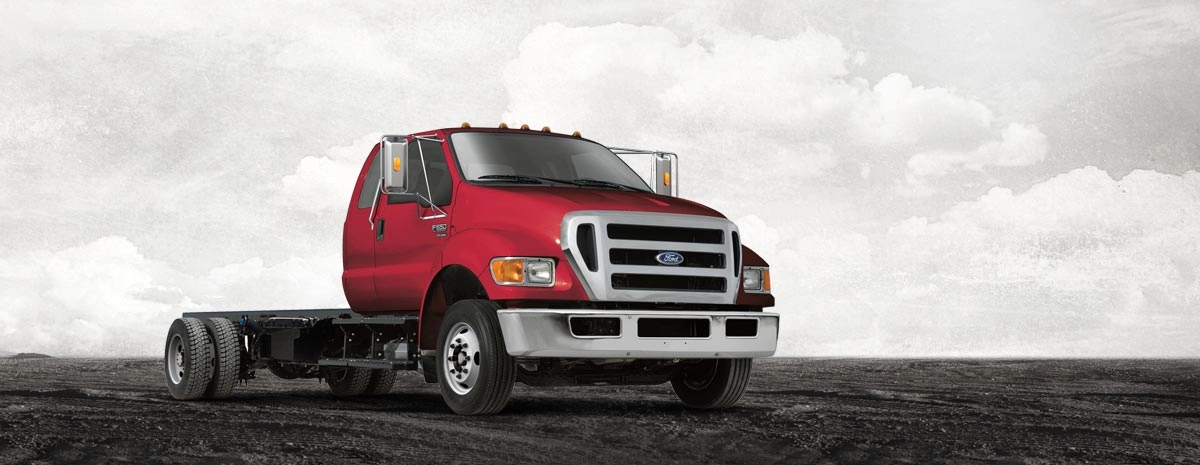 Red Ford F-650/F-750 medium-duty chassis cab truck with bare frame rails and no upfitted body, shown in a studio-style promotional photo against a cloudy sky backdrop