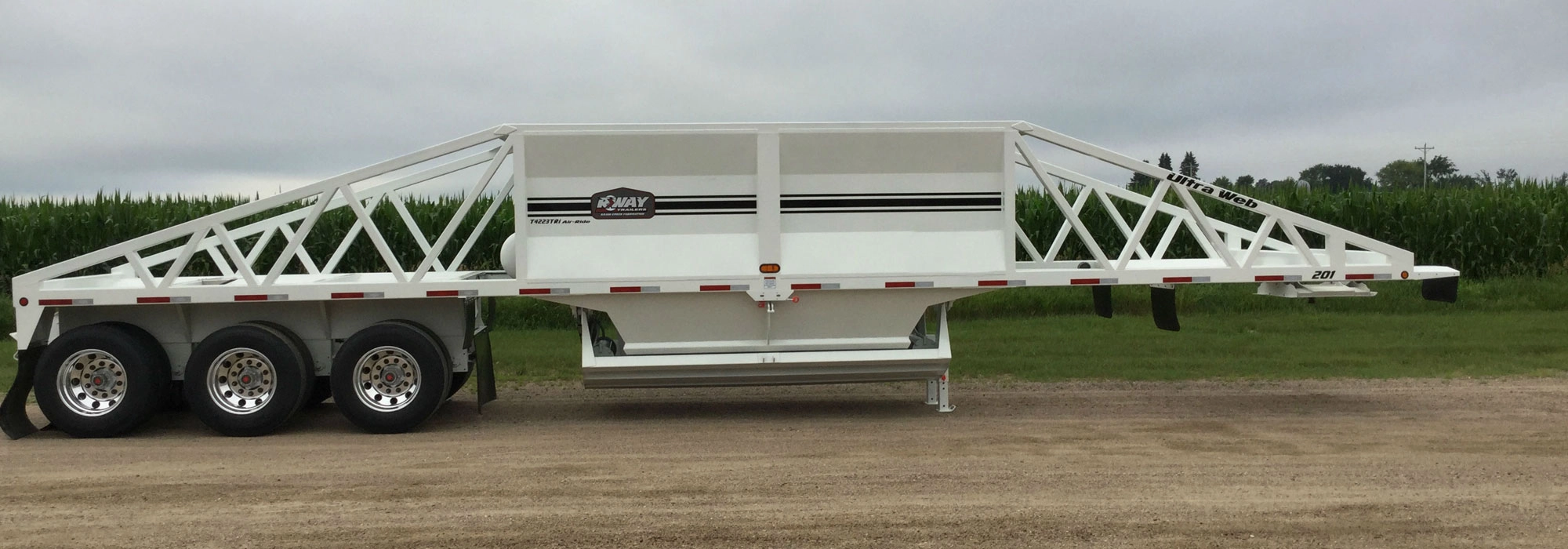 White tri-axle R-WAY bottom dump trailer with a truss-style side frame parked on a gravel lane beside a green cornfield