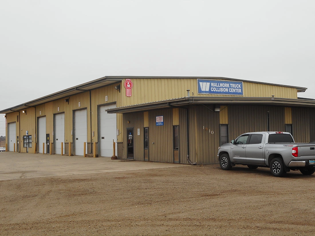 Exterior of the Wallwork Truck Collision Center building in Bismarck, North Dakota, a tan metal shop with multiple service bay doors, a Kenworth logo, and a pickup truck parked out front on a gravel lot