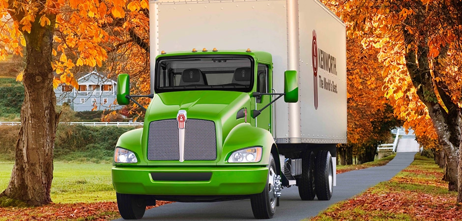 A green Kenworth T270 straight truck with a box body, driving down a tree-lined country lane with vivid autumn foliage and a farmhouse visible in the background.