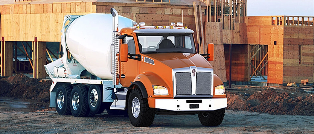 An orange Kenworth T880 with a white concrete mixer drum mounted on a tridem rear axle chassis, parked at a residential construction site with framed houses under construction in the background.