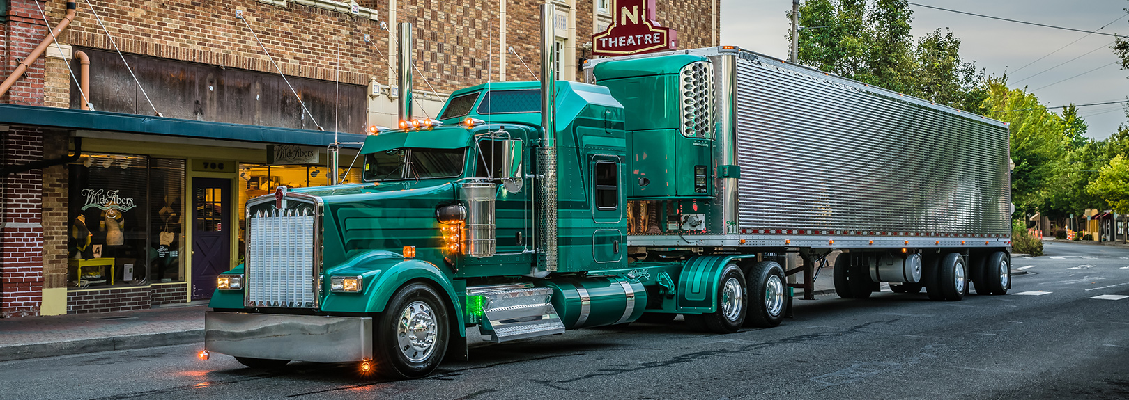 A classic green Kenworth W900L semi-truck with a chrome grille and dual exhaust stacks, pulling a silver reefer trailer, parked on a small-town street lined with brick storefronts and a lit theater marquee.